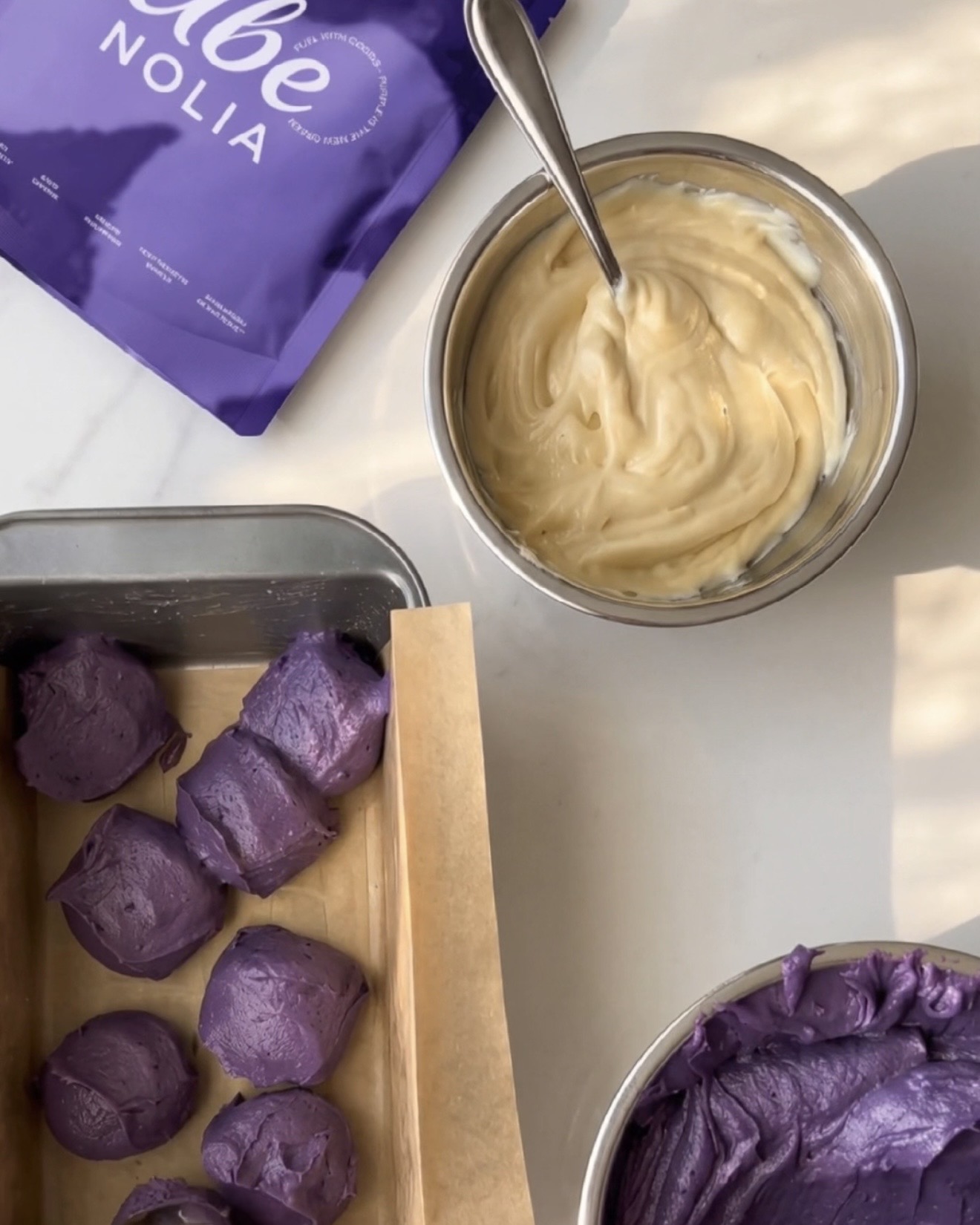 Valmis kotitekoinen ube halaya, vaniljataikina ja kauhallisia violettia ube-taikinaa leipävuoassa Ube Nolia ubejauhe -pussin vieressä