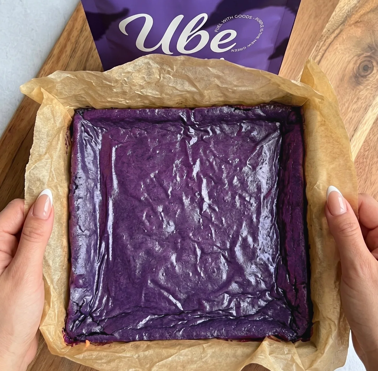 Syvän purppuraa ube-brownietaikinaa kerrostettuna leivinpaperilla vuorattuun neliövuokaan Ube Nolia -jauhepussin vieressä